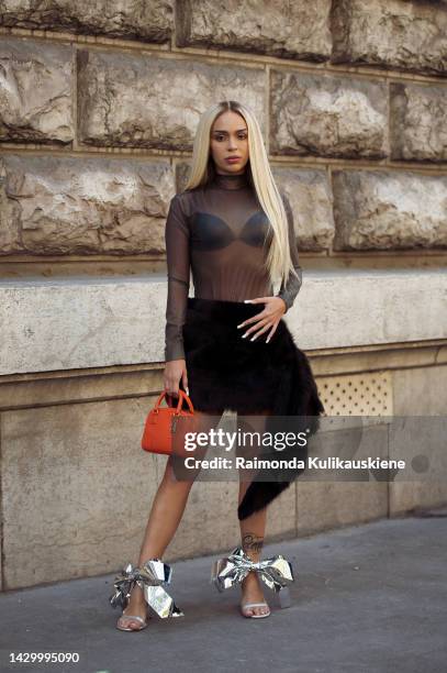 Ava Max wearing a black bikini, dark grey semtransparent long sleeve top, black fur skirt, silver metal look shoes with bows, and orange bag outside...