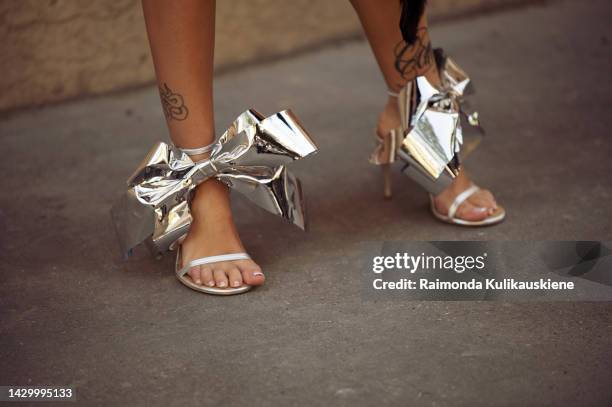 Ava Max wearing a black bikini, dark grey semtransparent long sleeve top, black fur skirt, silver metal look shoes with bows, and orange bag outside...