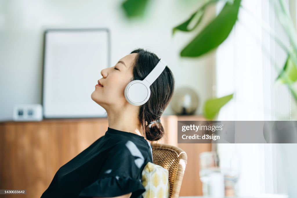 Close up, side profile of young Asian woman with eyes closed listening to music over headphones while relaxing on rattan arm chair at home, with sunlight shining through the window. Music, lifestyle and technology
