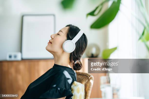 close up, side profile of young asian woman with eyes closed listening to music over headphones while relaxing on rattan arm chair at home, with sunlight shining through the window. music, lifestyle and technology - ヘッドフォン ストックフォトと画像