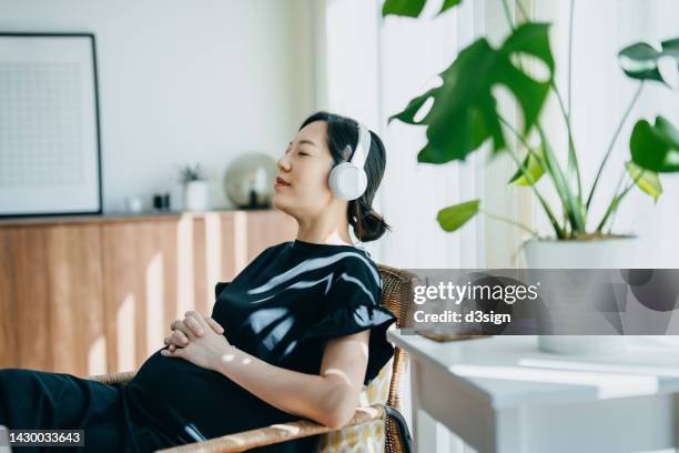 young asian pregnant woman with eyes closed listening to music over headphones while relaxing on rattan arm chair at home, with sunlight shining through the window. pregnancy lifestyle. pregnancy health and wellbeing - birthing chair stock pictures, royalty-free photos & images