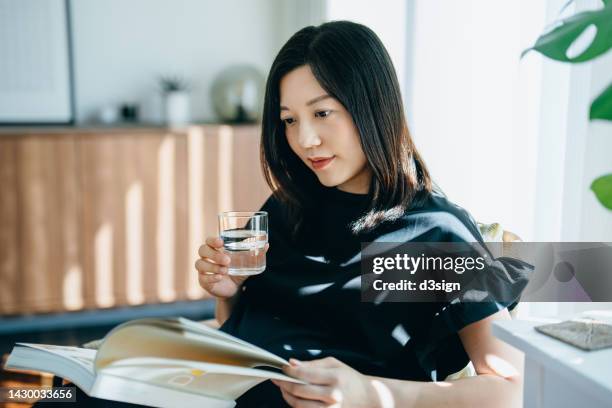 young asian woman reading a book while drinking a glass of water. enjoying a quiet time and relaxing environment against sunlight at cozy home - 女性　部屋 ストックフォトと画像