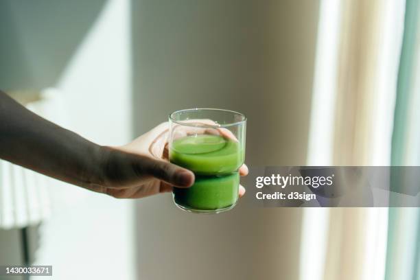 cropped hand of woman holding a glass of fresh green fruit and vegetable smoothie against white background with natural sunlight. healthy superfood. green colour, detox diet and healthy eating concept - juice foto e immagini stock