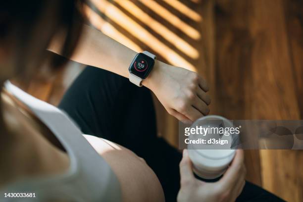 pregnant woman checking pulse and training performance on fitness tracker app on smartwatch after exercising at home. taking a rest and having a glass of milk after practicing yoga. pregnancy health and wellness. staying fit and healthy during pregnancy - staying indoors stock pictures, royalty-free photos & images