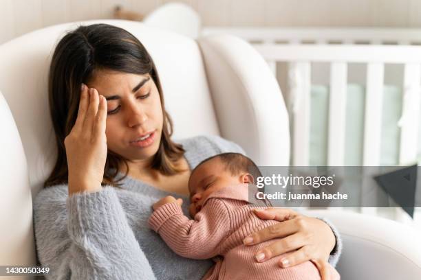 mother sitting on couch in nursery feeling postpartum depression with baby in her arms. - baby depression fotografías e imágenes de stock