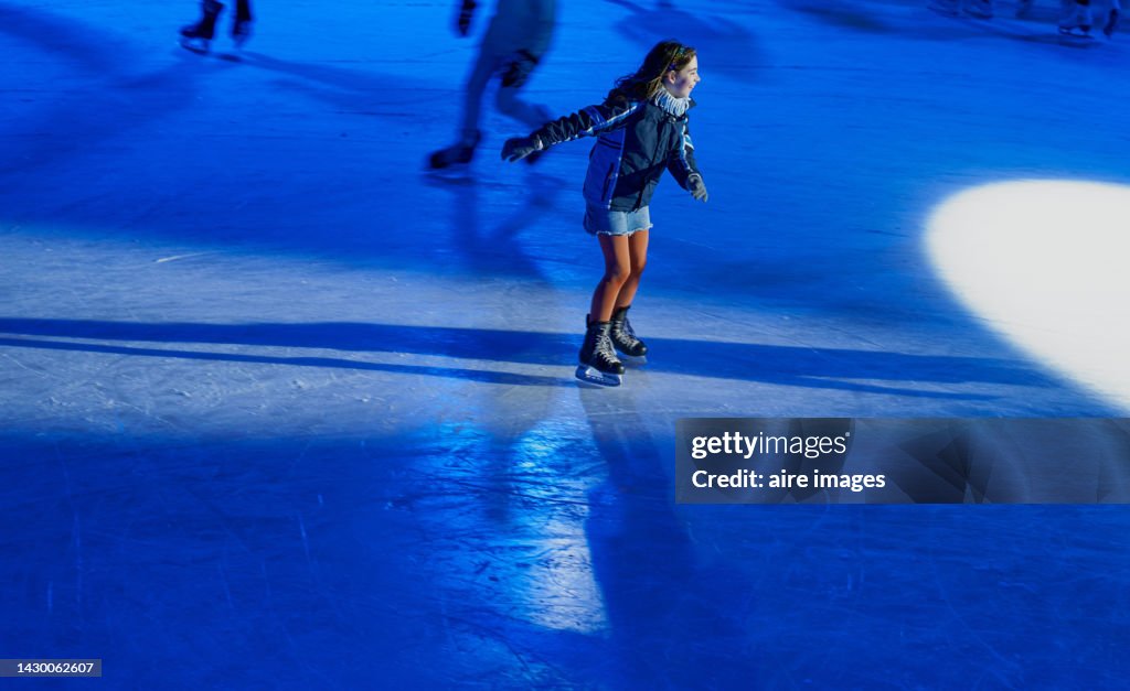 Caucasian teenage girl with long brown hair skating on an ice rink with skates, dressed in a skirt and jacket.