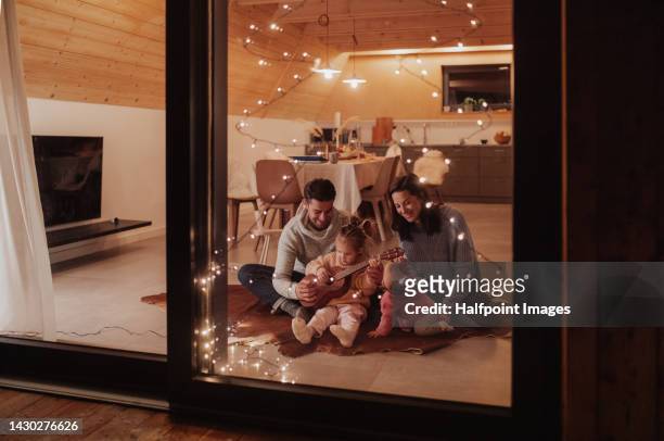 young cheerful family sitting in their living room and playing on guitar, enjoying christmas time. - low key beleuchtung stock-fotos und bilder