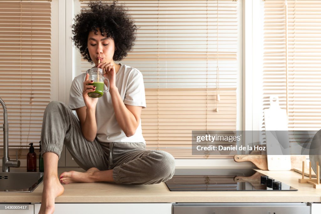 Mixed race black latina woman drinking healthy green juice sitting on kitchen counter. Copy space. Healthy eating.