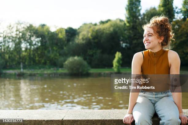 woman sat on wall next to river, smiling - locs hairstyle stockfoto's en -beelden