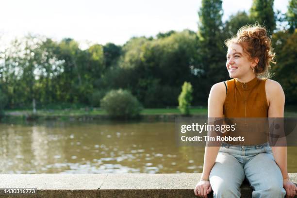 woman sat on wall next to river, smiling - candid curly hair stock pictures, royalty-free photos & images