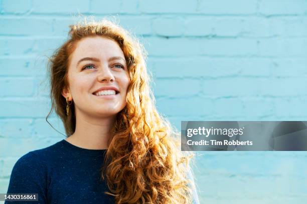 woman looking up, smiling next to blue back wall - cheveux roux photos et images de collection