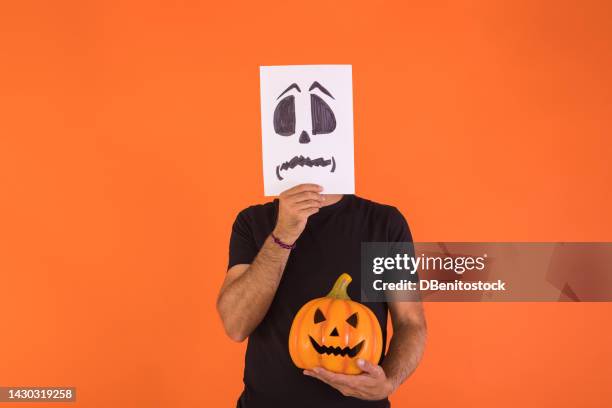 man covering his face with a paper on which a scared halloween face is painted, holding a pumpkin, on an orange background. concept of celebration, day of the dead and carnival. - cover monster face stock pictures, royalty-free photos & images