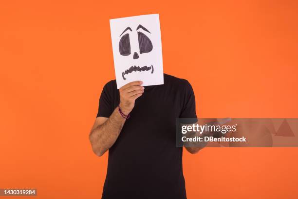 man covering his face with a paper on which a scared halloween face is painted, making a puzzled gesture, on an orange background. concept of celebration, day of the dead and carnival. - cover monster face stock pictures, royalty-free photos & images