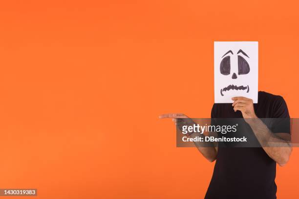 man covered his face with a paper on which a scared halloween face is painted, pointing a finger to the side, on an orange background. concept of celebration, day of the dead and carnival. - cover monster face stock pictures, royalty-free photos & images