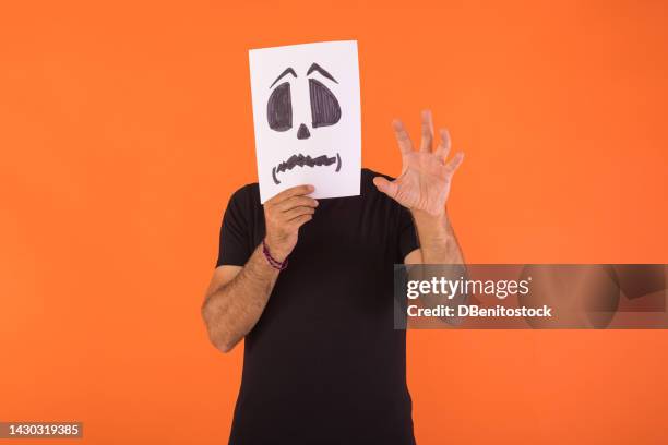 man covered his face with a paper on which a scared halloween face is painted, scaring with his hand, on an orange background. concept of celebration, day of the dead and carnival. - cover monster face stock pictures, royalty-free photos & images