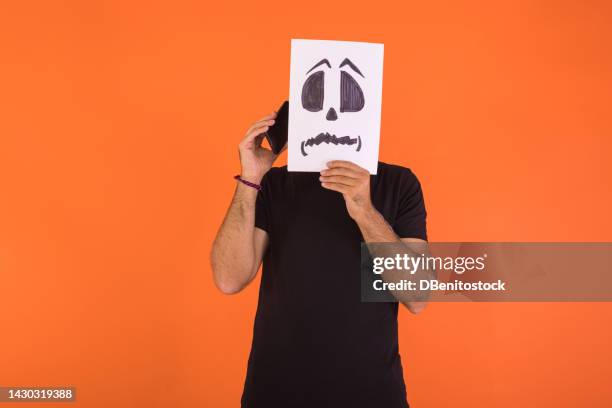 man covering his face with a paper on which a scared halloween face is painted, talking with the mobile phone, on an orange background. concept of celebration, day of the dead and carnival. - cover monster face stock pictures, royalty-free photos & images