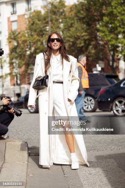 Chloe Harrouche wears black sunglasses, a white oversized long coat, a white shirt, a thin black leather belt, white Bermuda shorts, a black shiny...