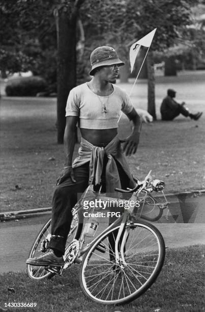 An unspecified Black man wearing a cropped t-shirt and a bucket hat, and a jacket tied around his waist, standing astride a racing cycle, location...