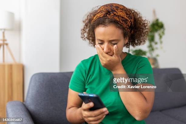 frustrated young adult woman reading message on smartphone. upset teenager student girl sitting on sofa holding mobile phone with hand on face. bad news, negative emotions, bad feelings concept - cyberbullying stockfoto's en -beelden