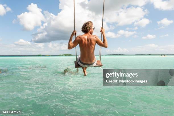 joven divirtiéndose en columpio en una hermosa laguna en un día soleado - columpiarse fotografías e imágenes de stock