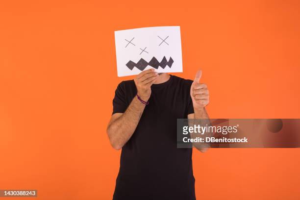man covering his face with a paper on which a scary smiling halloween face is painted, raising his thumb, on orange background. concept of celebration, day of the dead and carnival. - cover monster face stock pictures, royalty-free photos & images