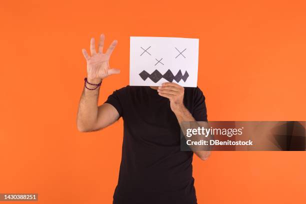 man covered his face with a paper on which a scared halloween face is painted, scaring with his hand, on an orange background. concept of celebration, day of the dead and carnival. - cover monster face stock pictures, royalty-free photos & images