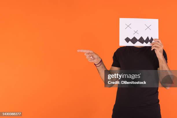 man covering his face with a paper on which a scary smiling halloween face is painted, pointing to the side, on orange background. concept of celebration, day of the dead and carnival. - cover monster face stock pictures, royalty-free photos & images