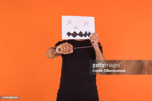 man covering his face with a paper on which a halloween jack o lantern pumpkin is painted, holding a knife, on an orange background. concept of celebration, day of the dead and carnival. - cover monster face stock pictures, royalty-free photos & images