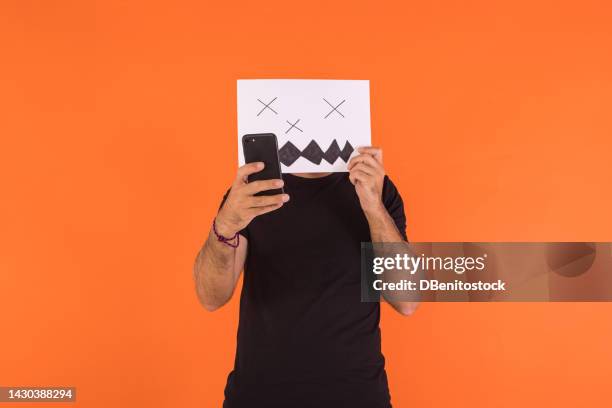 man covering his face with a paper on which a scared halloween face is painted, looking at the mobile phone, on an orange background. concept of celebration, day of the dead and carnival. - cover monster face stock pictures, royalty-free photos & images