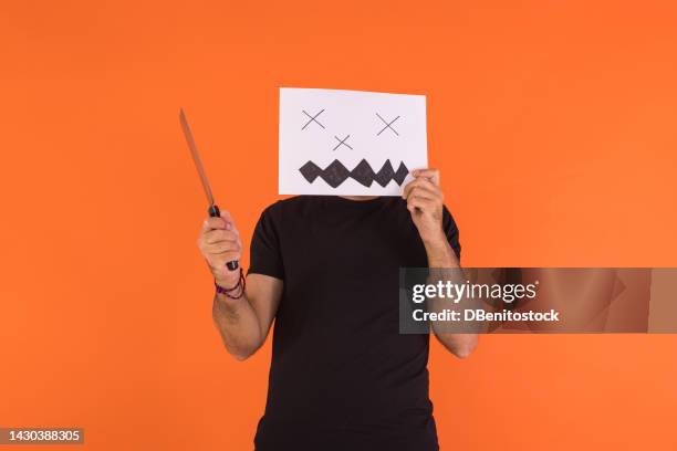 man covering his face with a paper on which a halloween jack o lantern pumpkin is painted, holding a knife, on an orange background. concept of celebration, day of the dead and carnival. - cover monster face stock pictures, royalty-free photos & images