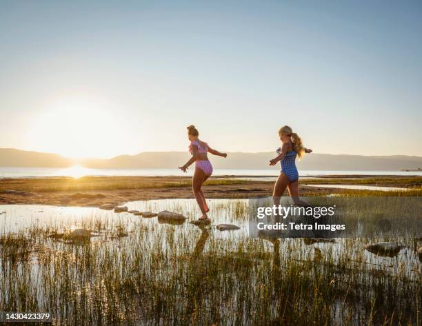 sisters (12-13, 14-15) walking on stepping stones in lake at sunrise - swimming shorts stock pictures, royalty-free photos & images