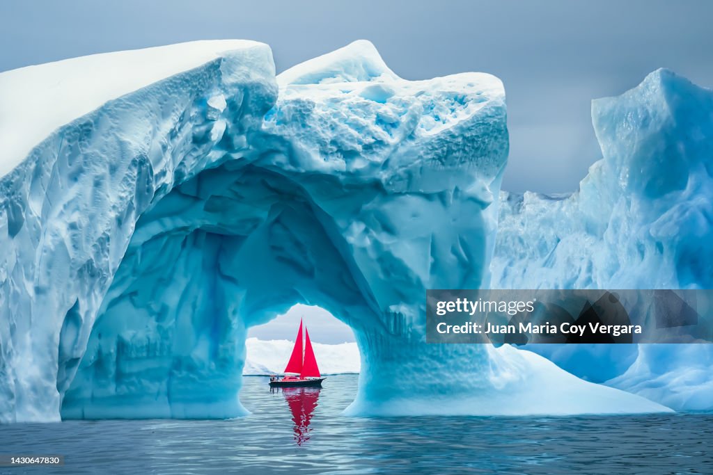 Red sailboat sailing under a majestic iceberg arch on sunny blue Artic Ocean in Greenland, Ilulissat Icefjord, Ilulissat, Disko Bay, Unesco World Heritage Site