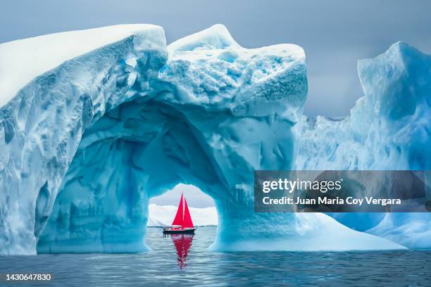 red sailboat sailing under a majestic iceberg arch on sunny blue artic ocean in greenland, ilulissat icefjord, ilulissat, disko bay, unesco world heritage site - disko bay stock pictures, royalty-free photos & images