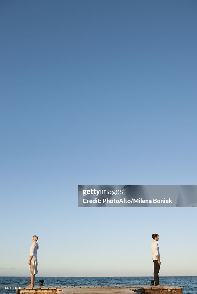 Couple standing back to back on pier, side view