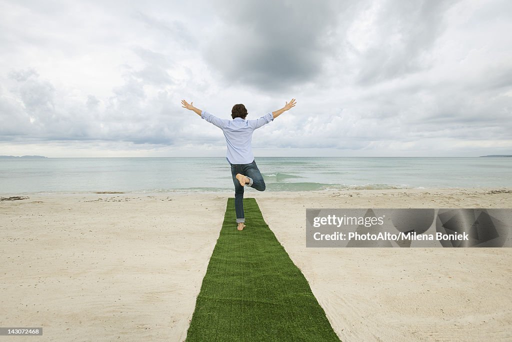 Man standing on one leg at end of carpet on beach with arms outstretched, rear view