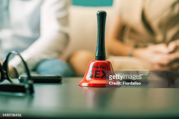 two mobile phones and a red bell with the inscription ring for sex, lying on a blue-green round sofa table. defocused loving couple in the background. family relationships in a couple, lack of time for personal communication, gadget addiction - bell stock pictures, royalty-free photos & images