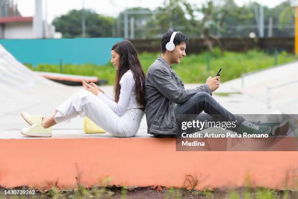 mujer joven y hombre joven sentados espalda con espalda usando sus teléfonos celulares - recostarse fotografías e imágenes de stock