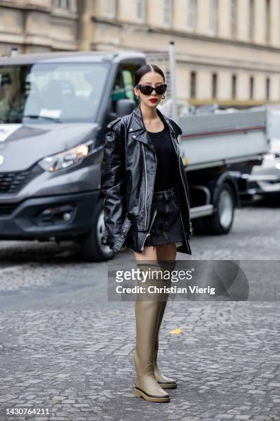 Irene Kim wears black varnished leather jacket, denim mini skirt, khaki over knee rain boots Chanel, black top outside Sacai during Paris Fashion...