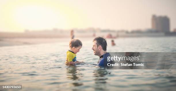 father and infant son having fun at the beach in the shallow seaside water at sunset - united arab emirates tourist stock pictures, royalty-free photos & images