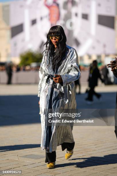 Naomi Campbell wears yellow futurist sunglasses from Louis Vuitton, a gray and silver shiny fabric long coat from Louis Vuitton, colored pearls...