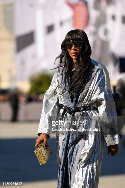 Naomi Campbell wears yellow futurist sunglasses from Louis Vuitton, a gray and silver shiny fabric long coat from Louis Vuitton, colored pearls...