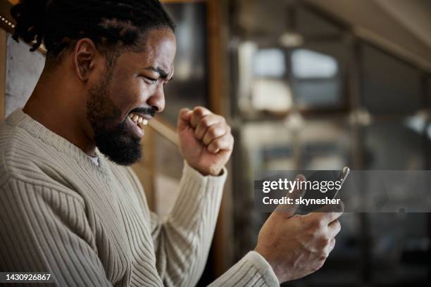 cheerful black man celebrating success while using cell phone at home. - punching the air stock pictures, royalty-free photos & images