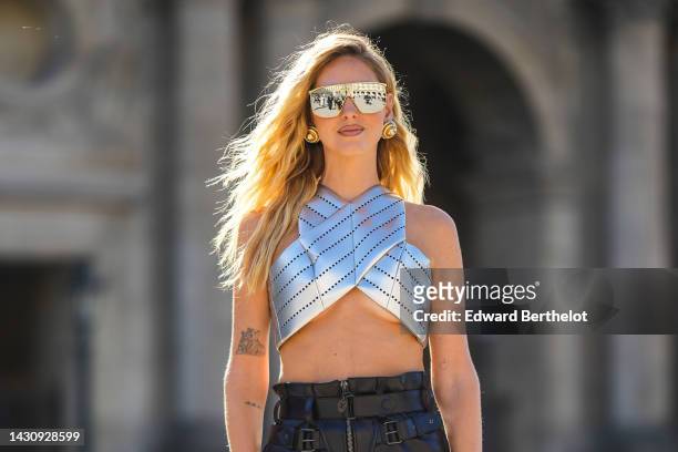 Chiara Ferragni wears black futurist sunglasses, gold oversized earrings, a silver shiny leather cut-out stripe pattern / halter neck strap cropped...