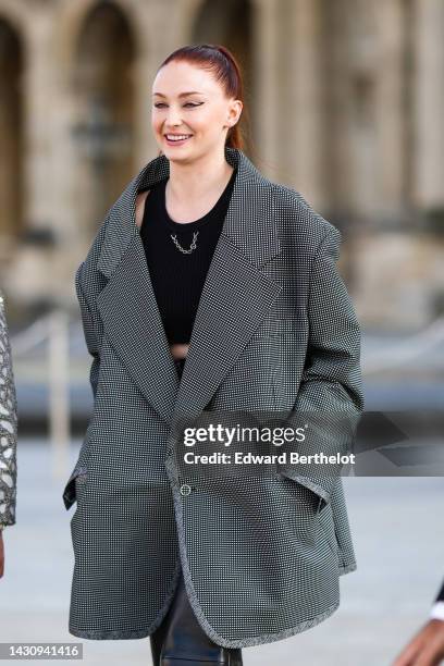 Sophie Turner wears a black cropped tank-top with a silver chain necklace, a black and white small checkered print pattern oversized jacket, black...