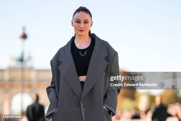 Sophie Turner wears a black cropped tank-top with a silver chain necklace, a black and white small checkered print pattern oversized jacket, black...