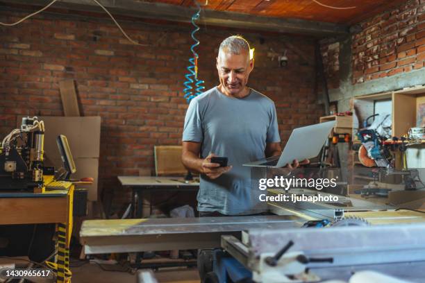 smiling carpenter using smartphone in carpentry workshop. - laptop stand stock pictures, royalty-free photos & images