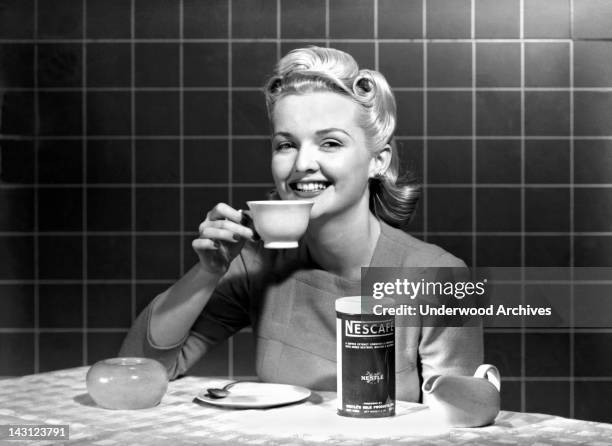 Single woman at a kitchen table enjoys a cup of Nescafe, circa 1960.