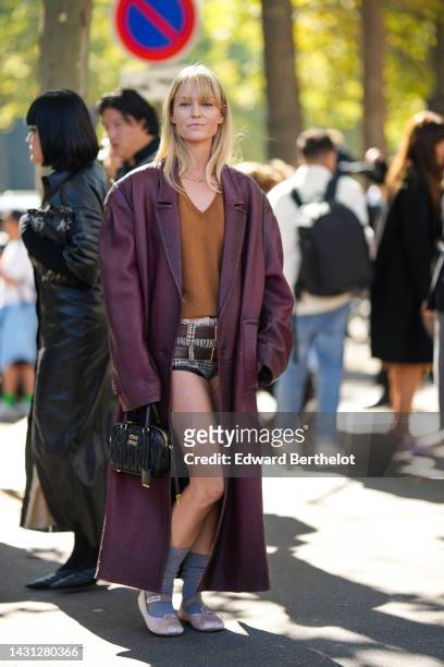 Jeanette Madsen wears a gold necklace, a brown V-neck / wool pullover, a burgundy shiny leather oversized long coat, dark brown shiny leather belts,...