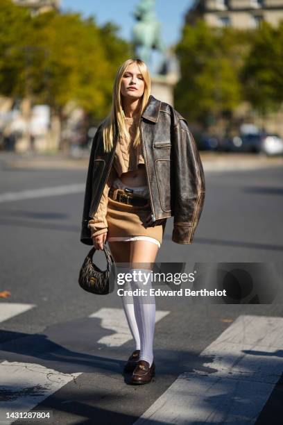 Camille Razat wears a beige braided wool cropped pullover, a dark brown shiny leather oversized jacket, a beige wool and white latte silk short skirt...