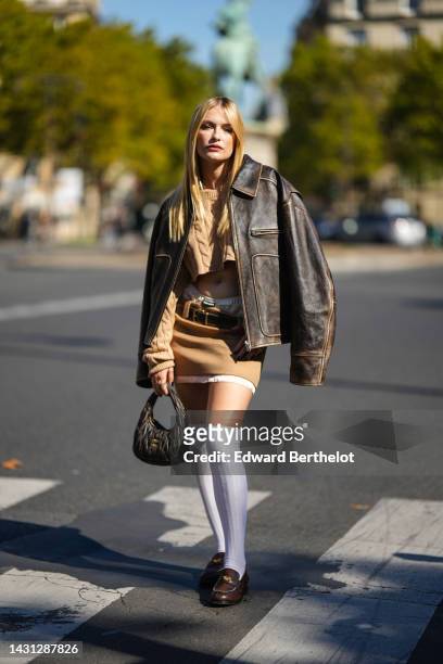 Camille Razat wears a beige braided wool cropped pullover, a dark brown shiny leather oversized jacket, a beige wool and white latte silk short skirt...